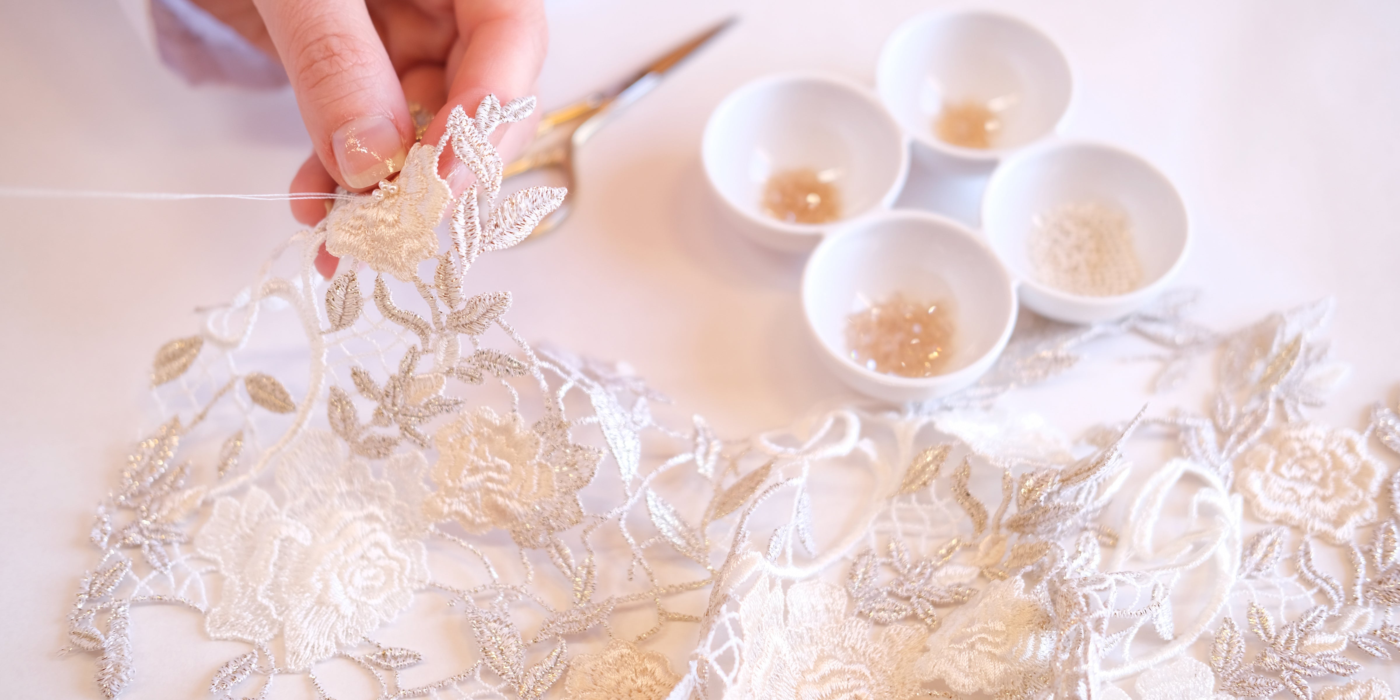 Close-up of hand embroidery in progress on metallic floral lace. A needle threads a pearl into place, with small ceramic bowls of sequins and beads nearby. The image captures the detailed artistry and delicate hand-finishing that defines bespoke, made-to-order bridal design.
