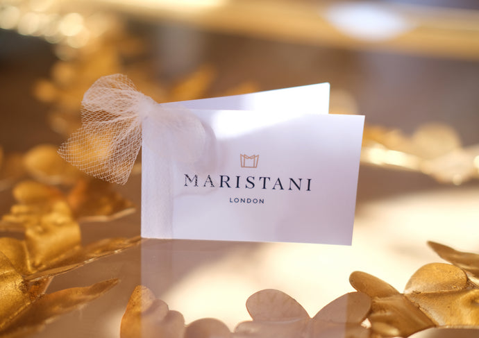 Close-up of a Maristani London brand card placed on a reflective surface adorned with golden floral accents. The white card features the Maristani logo in gold above the brand name in elegant black serif text, tied with a soft ivory tulle bow. Warm lighting enhances the romantic presentation.