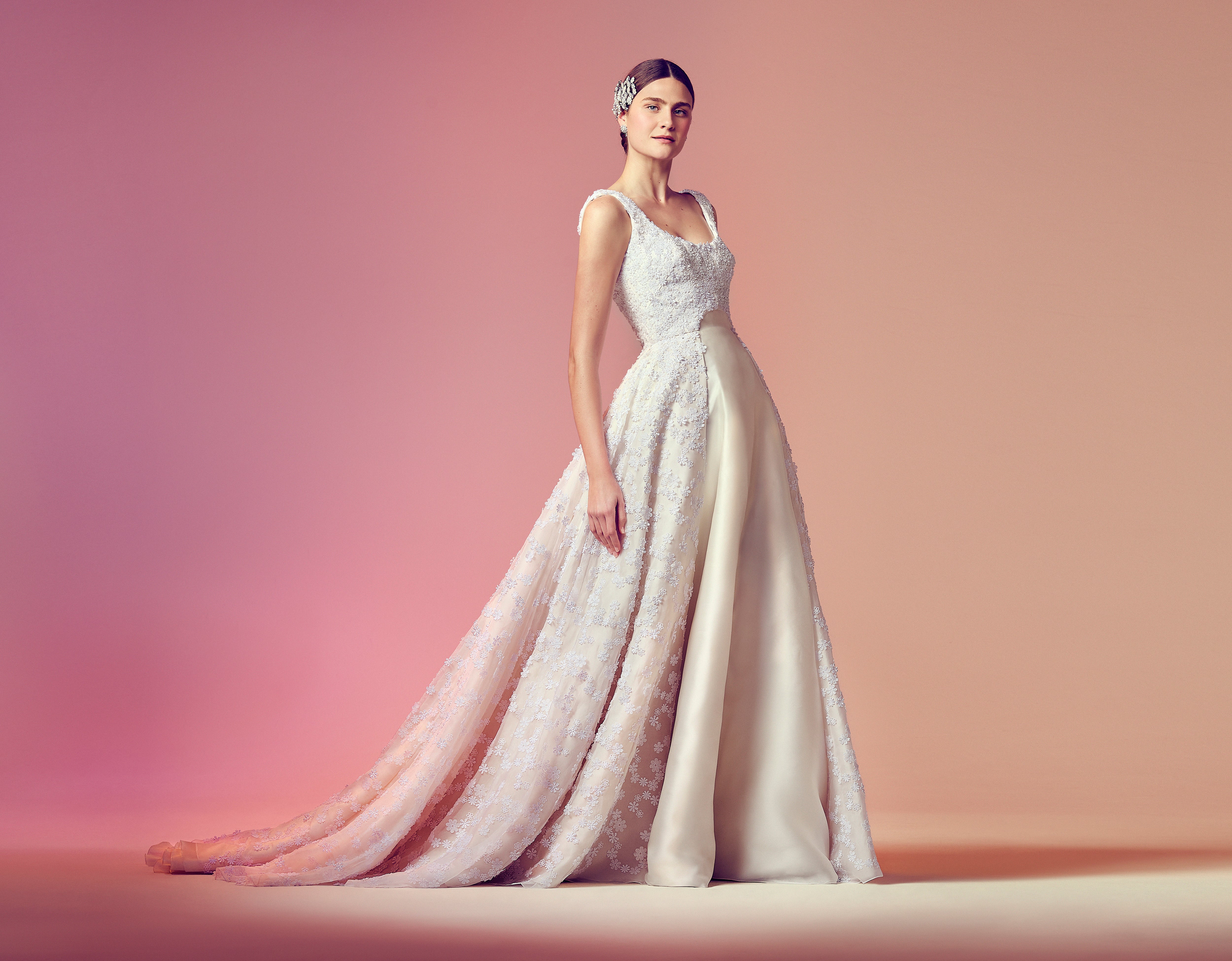Model wearing a custom, bespoke, made-to-order bridal gown with a scooped neckline and structured bodice. The dress features tonal floral appliqué over a full overskirt, revealing a smooth satin underlayer. Styled with a floral headpiece, set against a soft pink gradient backdrop.