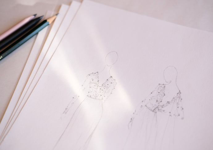 Close-up of bridal design sketches on white paper, featuring two pencil-drawn figures in embellished gowns with sheer sleeves and fitted bodices. Soft sunlight casts shadows across the page. A few coloured pencils lie beside the stack, hinting at the bespoke design process behind each gown.