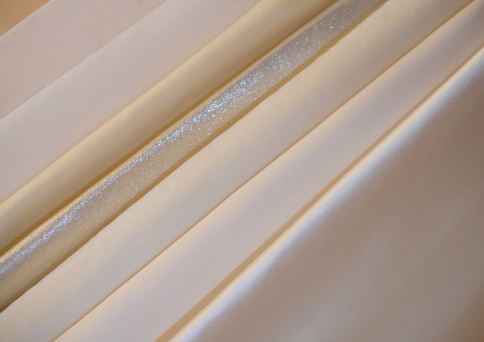 Close-up of a selection of custom bridal fabrics in soft ivory tones, including matte organza, shimmering glitter tulle, and satin with a smooth, light-reflective finish. The image captures the material options used in bespoke, made-to-order gown construction.