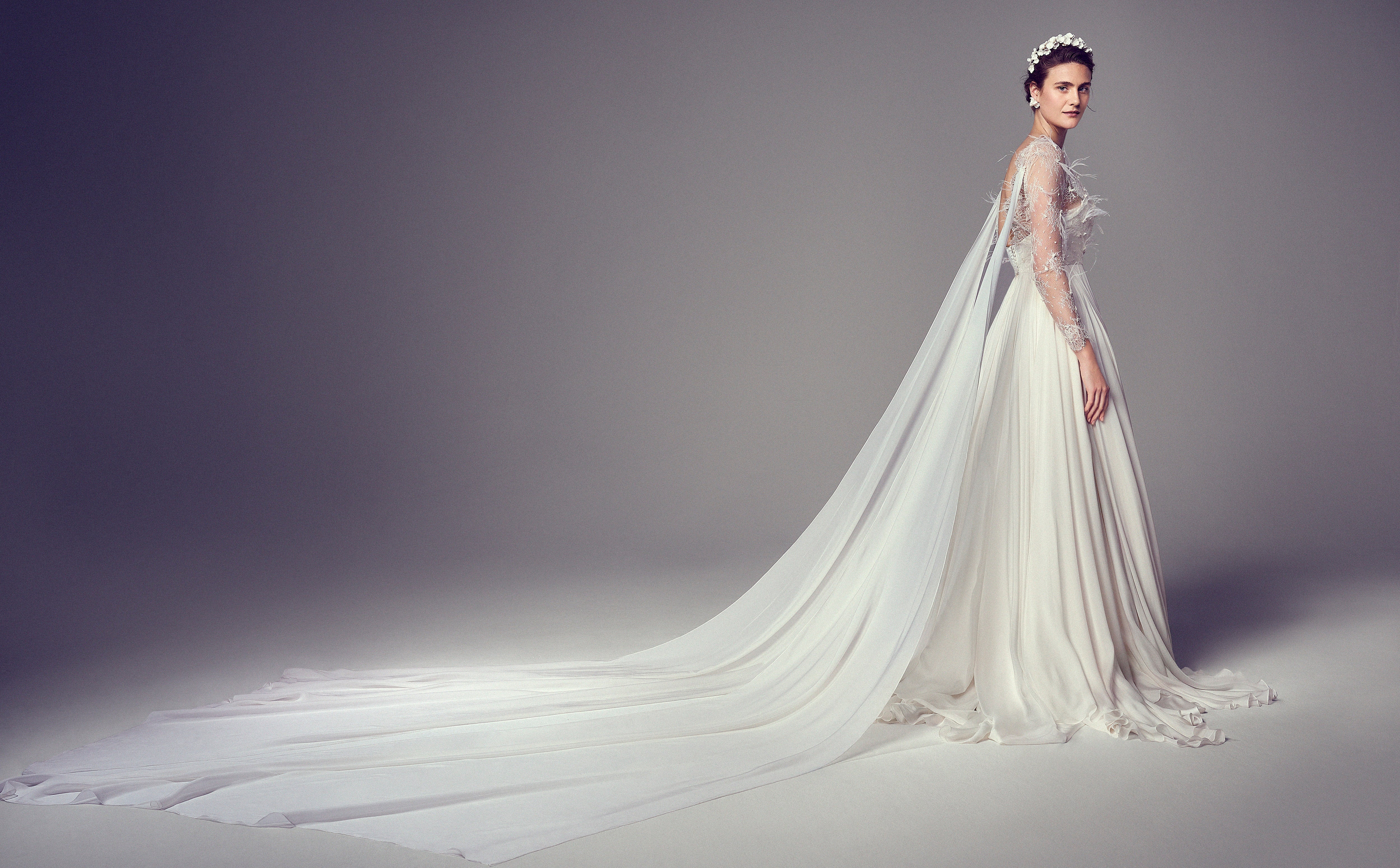 Model wears a floor-length gown with sheer embroidered sleeves and soft chiffon panels draping from the shoulders, creating an ethereal train.