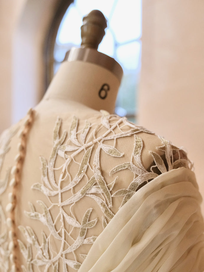 Close-up of a custom, bespoke, made-to-order bridal gown on a mannequin, showing intricate leaf embroidery in soft ivory and sage tones. The back features delicate covered buttons and sheer draping over one shoulder.