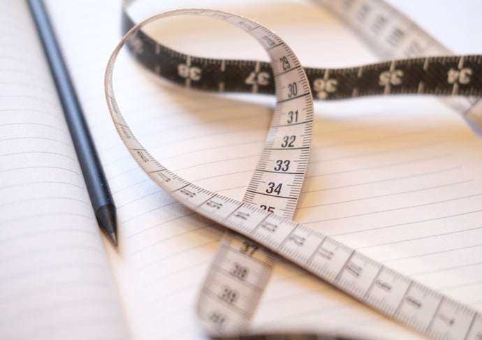 Flat lay of a black measuring tape coiled on a lined notepad, next to a sharpened pencil. Capturing the precision behind bespoke tailoring, the image reflects the beginning stages of custom gown creation, where each measurement informs fit and silhouette.