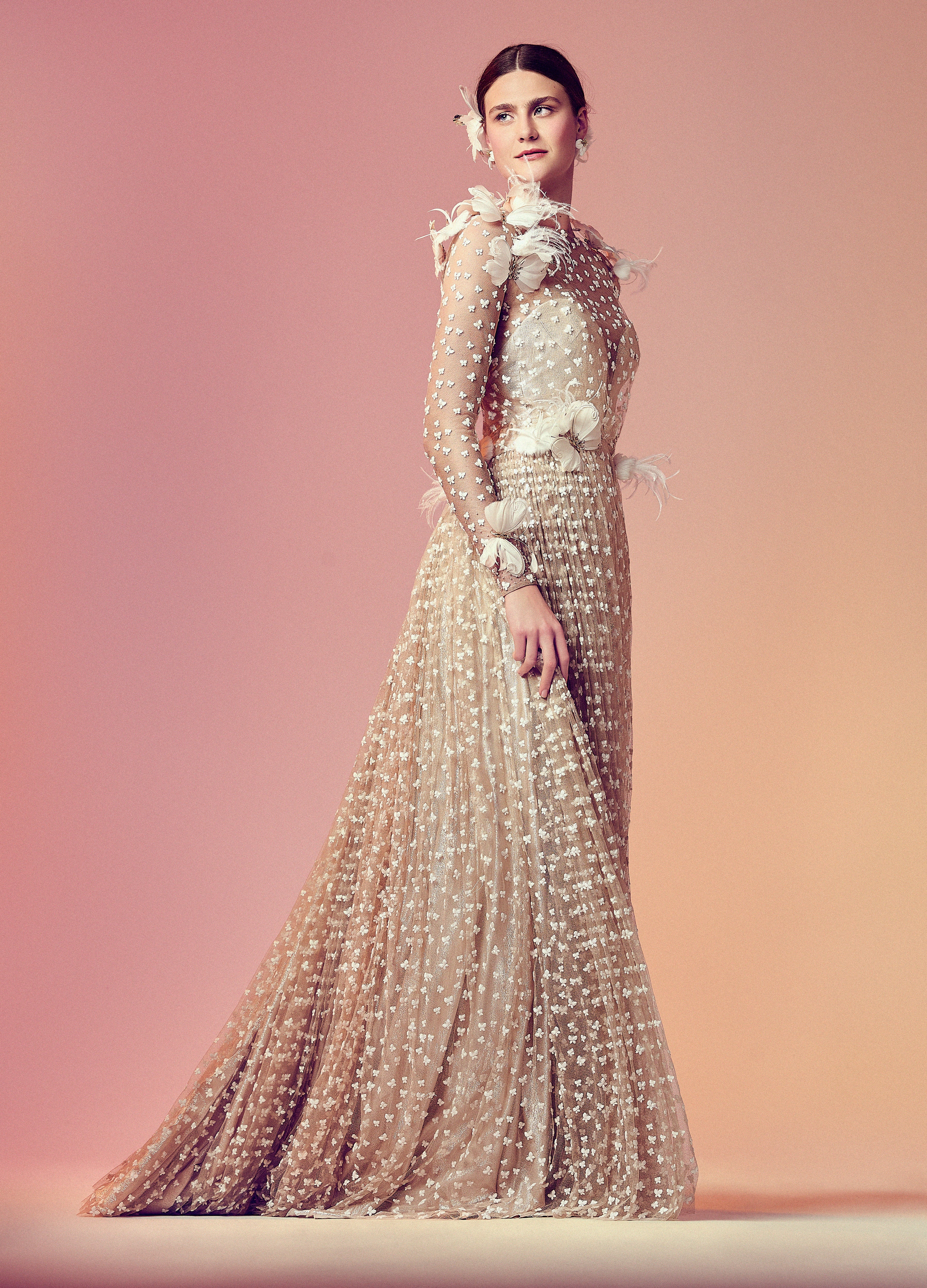 Model poses in a blush-toned tulle gown embroidered with butterflies and trimmed with feathers. The long sleeves and pleated skirt create a graceful silhouette, framed by soft texture and sparkle. Set against a warm pink-to-peach gradient backdrop.