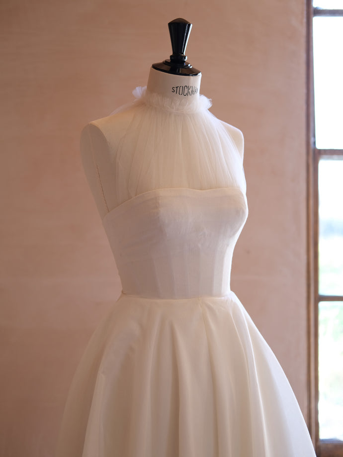 A custom, bespoke, made-to-order bridal gown in progress, displayed on a Stockman mannequin. The gown features a softly structured bodice with ruched tulle neckline and a voluminous skirt. The warm-toned studio backdrop evokes artisanal craftsmanship and quiet elegance.