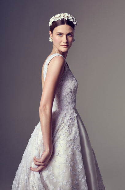 Model in a custom, bespoke, made-to-order bridal gown with a sleeveless silhouette and structured bodice, richly embroidered in tonal floral appliqué. The voluminous skirt flows with a smooth satin-like texture. Styled with a handcrafted floral headband and earrings, against a grey backdrop.