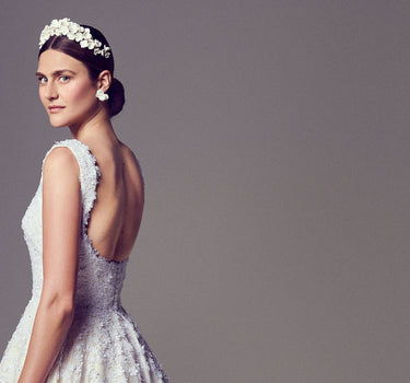 Model in a custom, bespoke, made-to-order bridal gown with an open back and sleeveless silhouette. The bodice and skirt feature three-dimensional floral embroidery that blends into the gown’s base. Styled with a handcrafted floral headband and earrings, against a grey backdrop.
