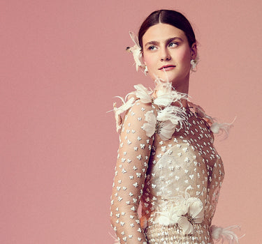 Model wears a sheer gown embroidered with delicate butterflies and accented with soft feathers. The transparent overlay reveals a shimmering base, with butterflies clustered at the shoulder and waist. Shot against a dusky pink backdrop.